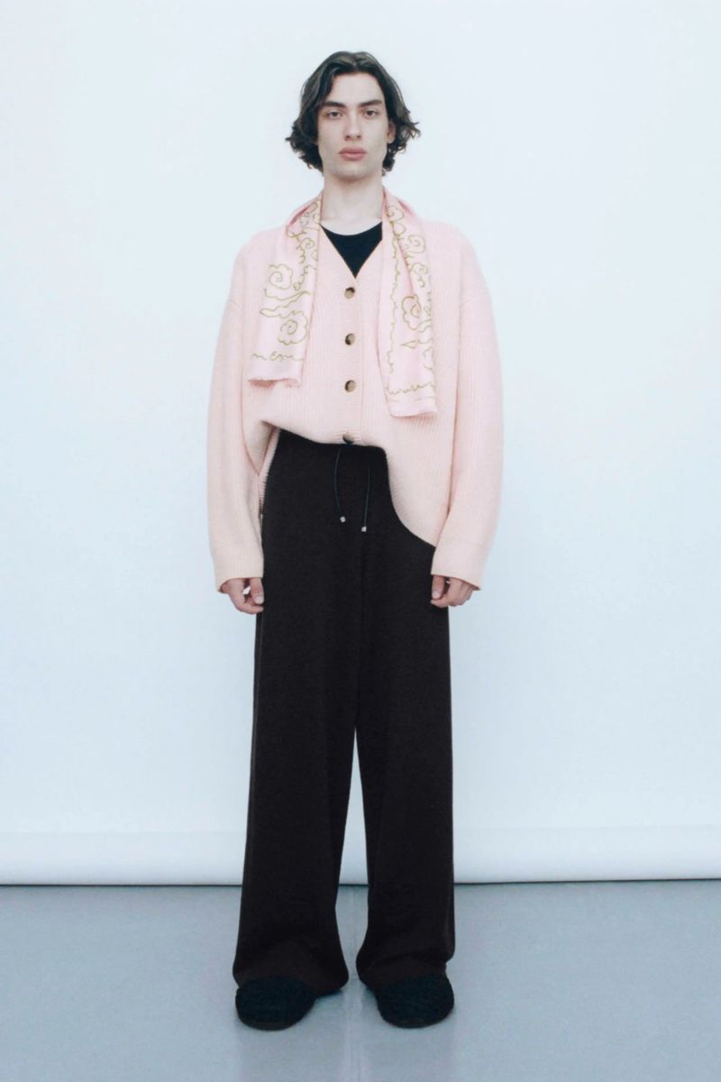 Full shot of a young adult man posing in a studio wearing a light pink cardigan with scarf, and brown pants, against a white backdrop.