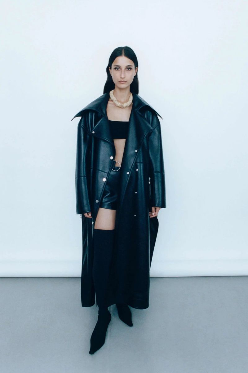 Fashionable woman poses in black leather trench coat, shorts, boots, and statement necklace against a neutral backdrop in studio lighting.