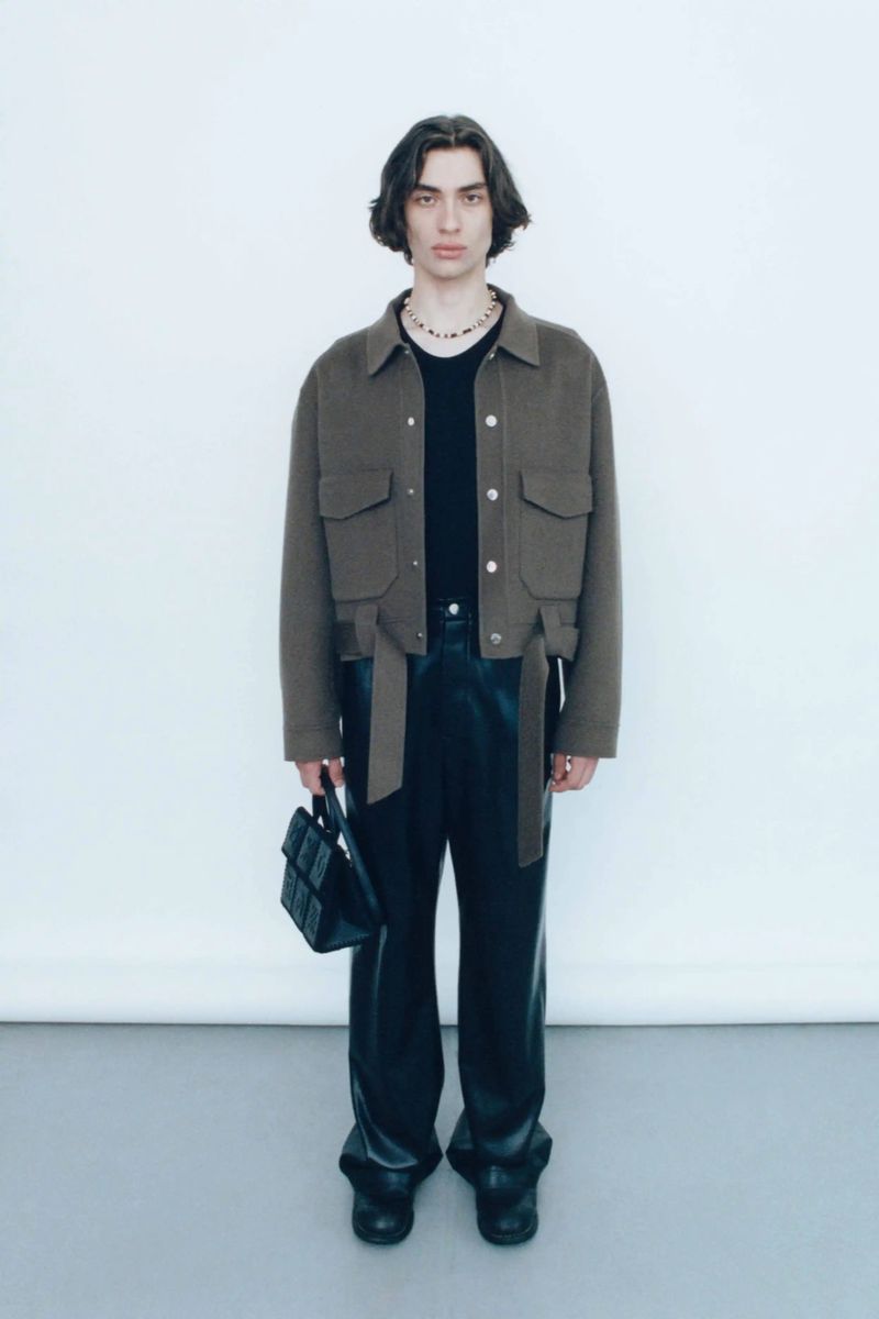 Full shot of a young adult man in a studio wearing a jacket, black shirt and black leather pants and holding a small black handbag.