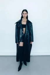 Swati Eck wears an oversized black leather trench coat, bandeau top, shorts, and pointed black boots against a white studio wall.