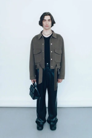 Model wearing a cropped brown utility jacket, black leather trousers, and holding a small black handbag against a white background.