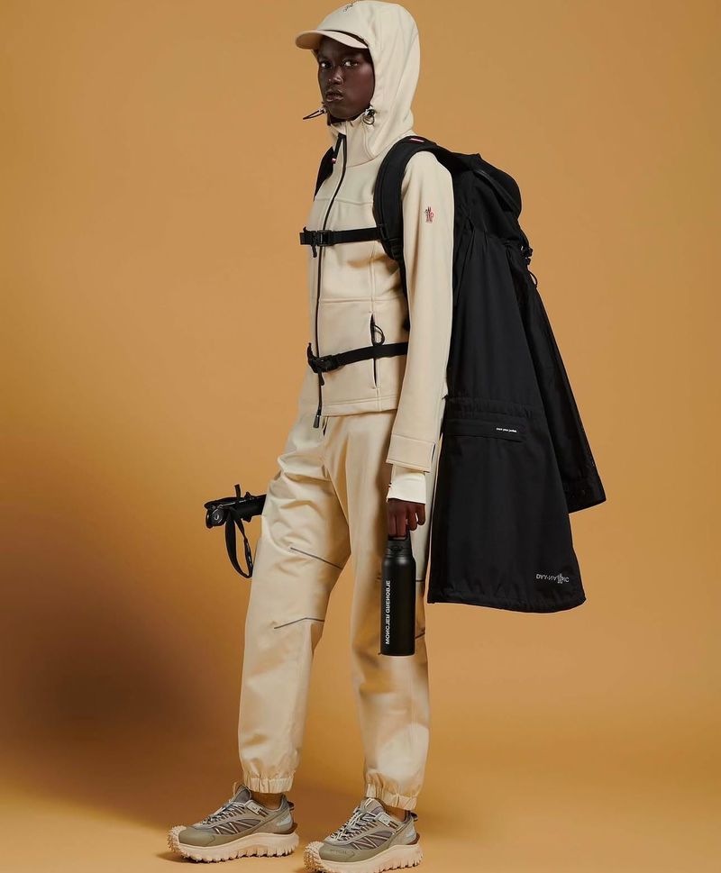 Dark-skinned male model in beige athleisure outfit with black backpack and water bottle, standing in a neutral studio.