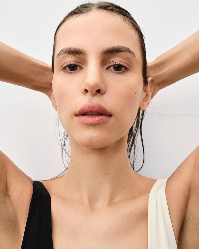 Close-up portrait of an adult female model with wet hair pulled back, hands behind her head, wearing a black and white top on a white backdrop.
