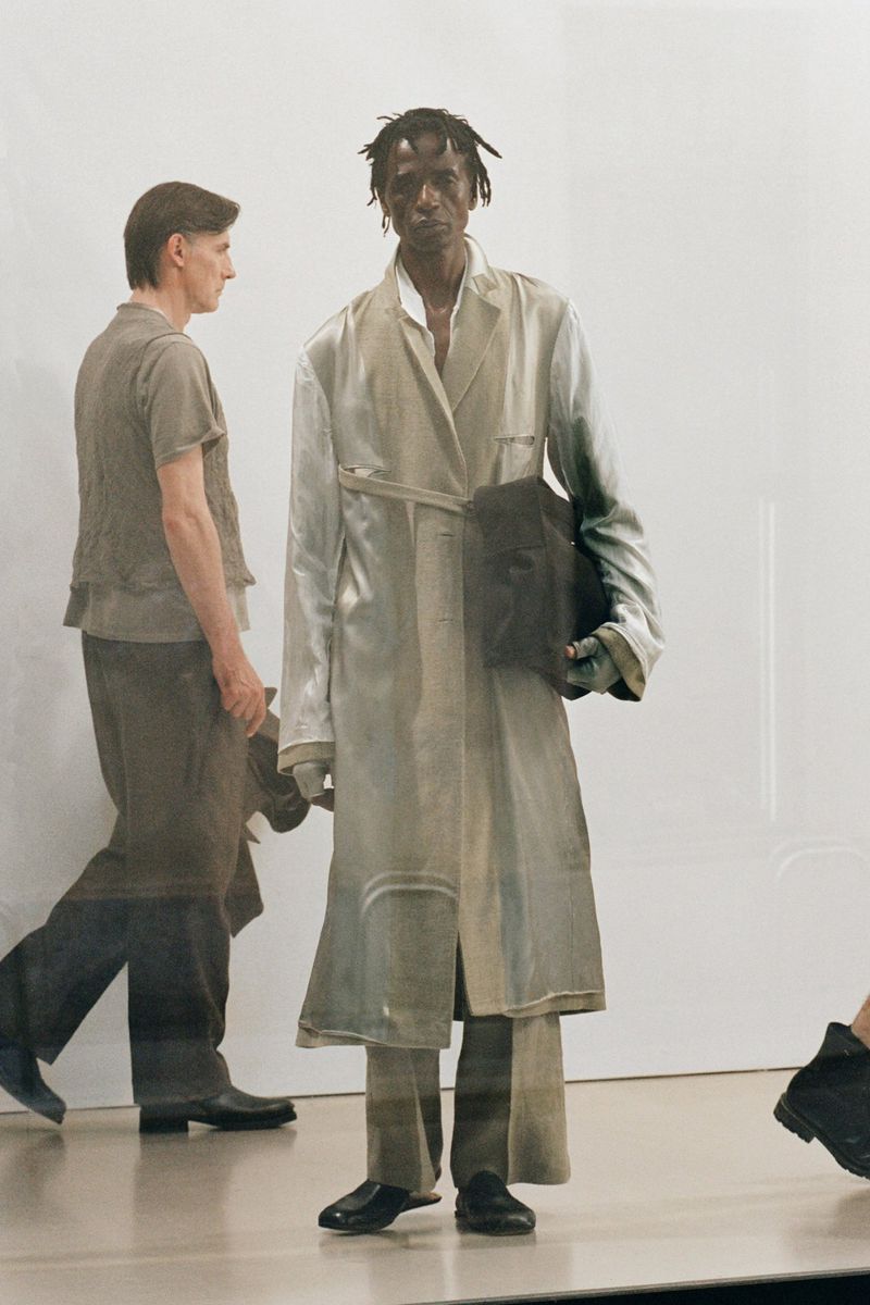 Runway fashion show with a man in a light trench coat holding a dark bag, other models partially visible, bright studio backdrop.