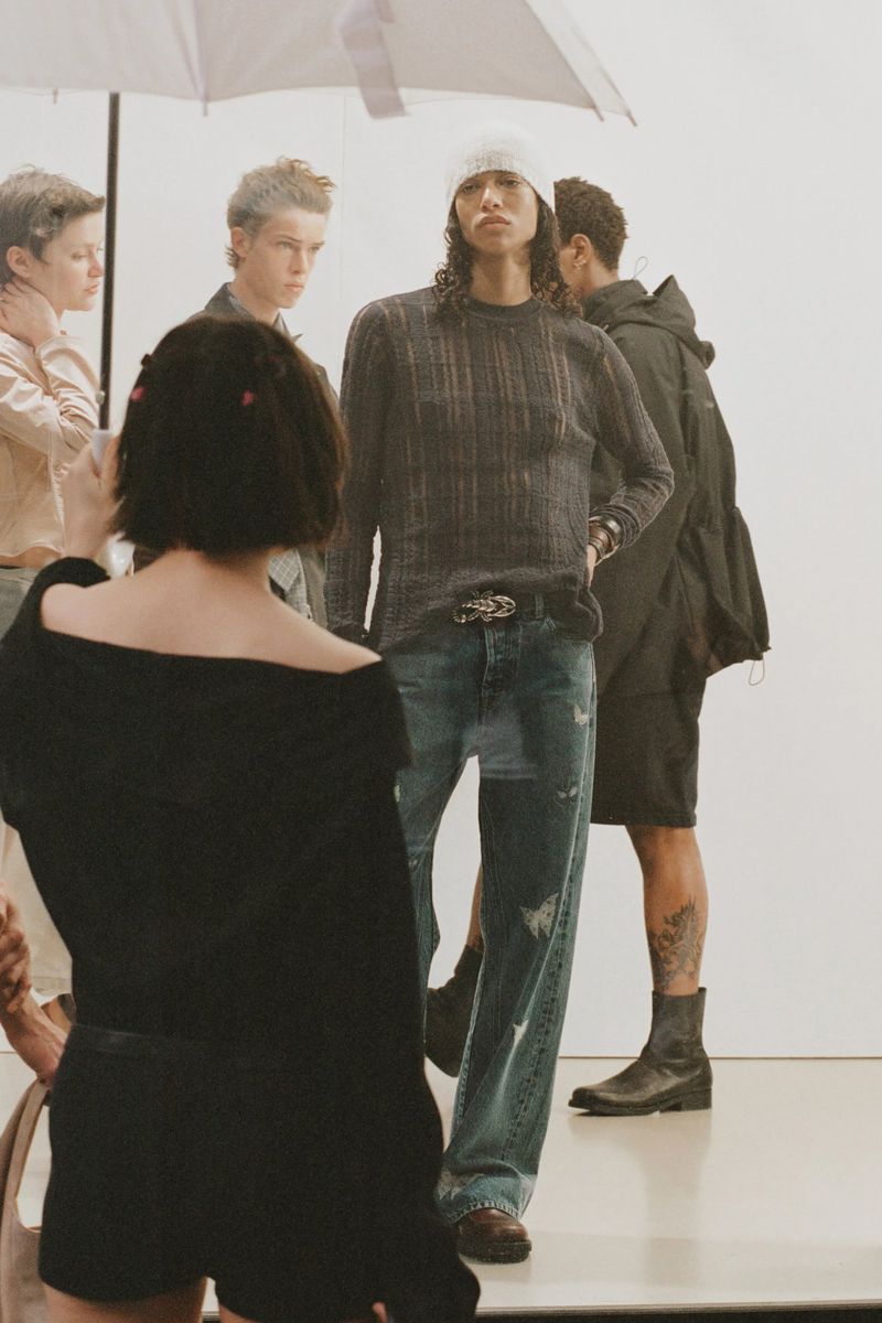 A group of young adult fashion models pose in a bright photo studio, wearing casual streetwear clothing like sweaters, jeans, and boots.
