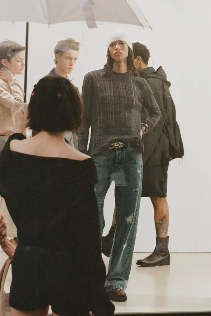 A group of young adult fashion models pose in a bright photo studio, wearing casual streetwear clothing like sweaters, jeans, and boots.