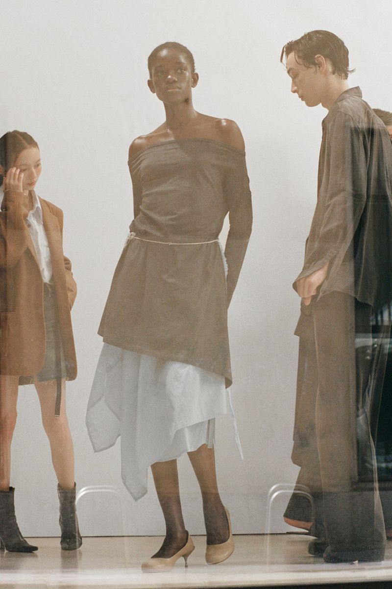 Three fashion models showcase neutral-toned clothing in a studio. The central model wears an off-shoulder top and layered skirt.