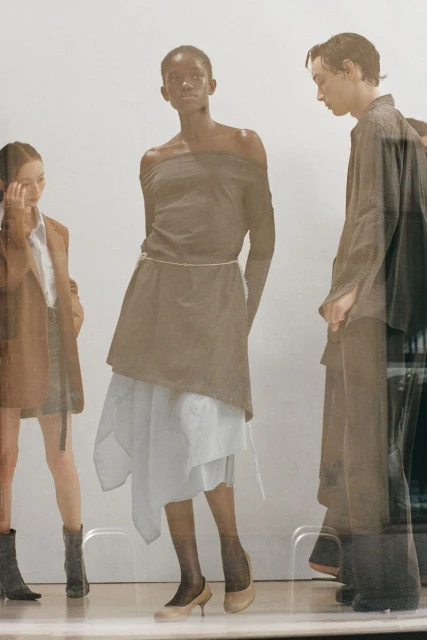 Three fashion models showcase neutral-toned clothing in a studio. The central model wears an off-shoulder top and layered skirt.