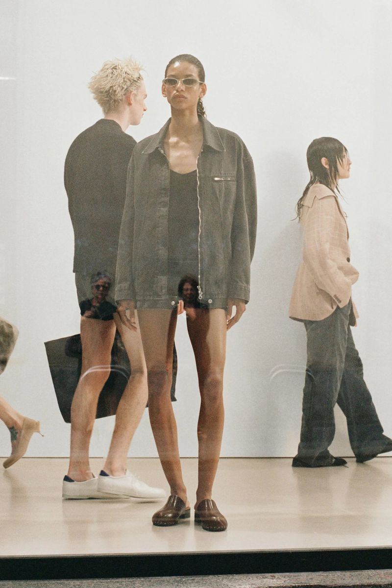 Fashion shot of a model in a grey jacket and brown shoes, with other people in the background, creating a layered, dynamic composition.