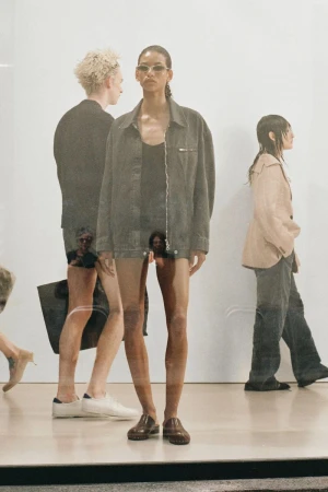 Fashion shot of a model in a grey jacket and brown shoes, with other people in the background, creating a layered, dynamic composition.