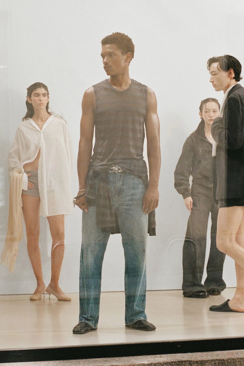 A group of male and female fashion models pose on a white floor in relaxed poses, wearing modern clothing with a clear fashion studio backdrop.