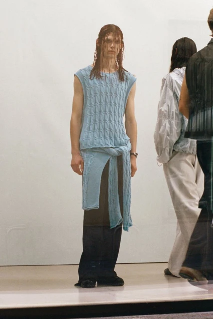 Runway fashion show featuring a male model in a light blue knit top, baggy black pants, and wet hair, with additional models in the background.