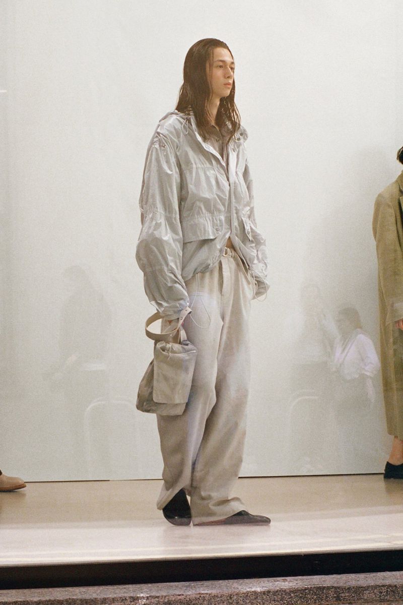 Adult male fashion model in a light grey windbreaker jacket and linen pants with a linen purse stands in a photo studio.