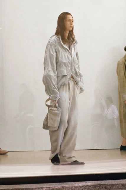 Adult male fashion model in a light grey windbreaker jacket and linen pants with a linen purse stands in a photo studio.
