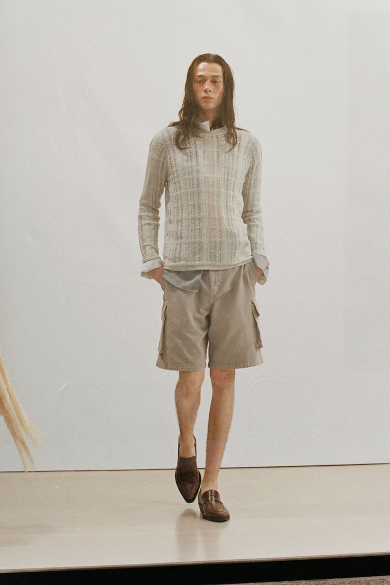 A long-haired person walks on a neutral surface, wearing a gray knit hoodie, tan shorts with side pockets, and brown leather loafers.