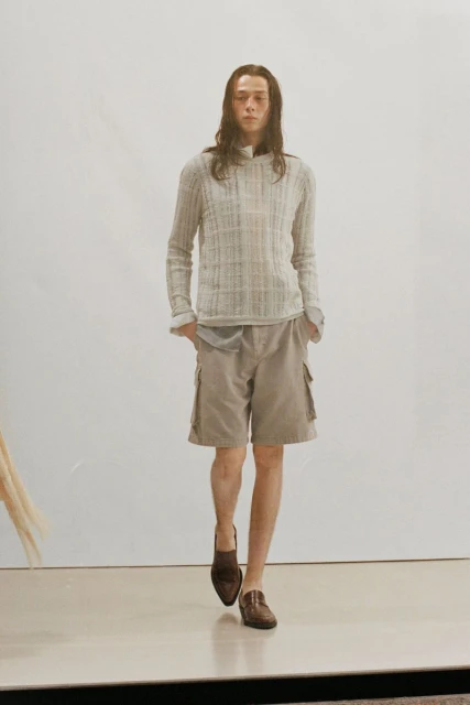 A long-haired person walks on a neutral surface, wearing a gray knit hoodie, tan shorts with side pockets, and brown leather loafers.
