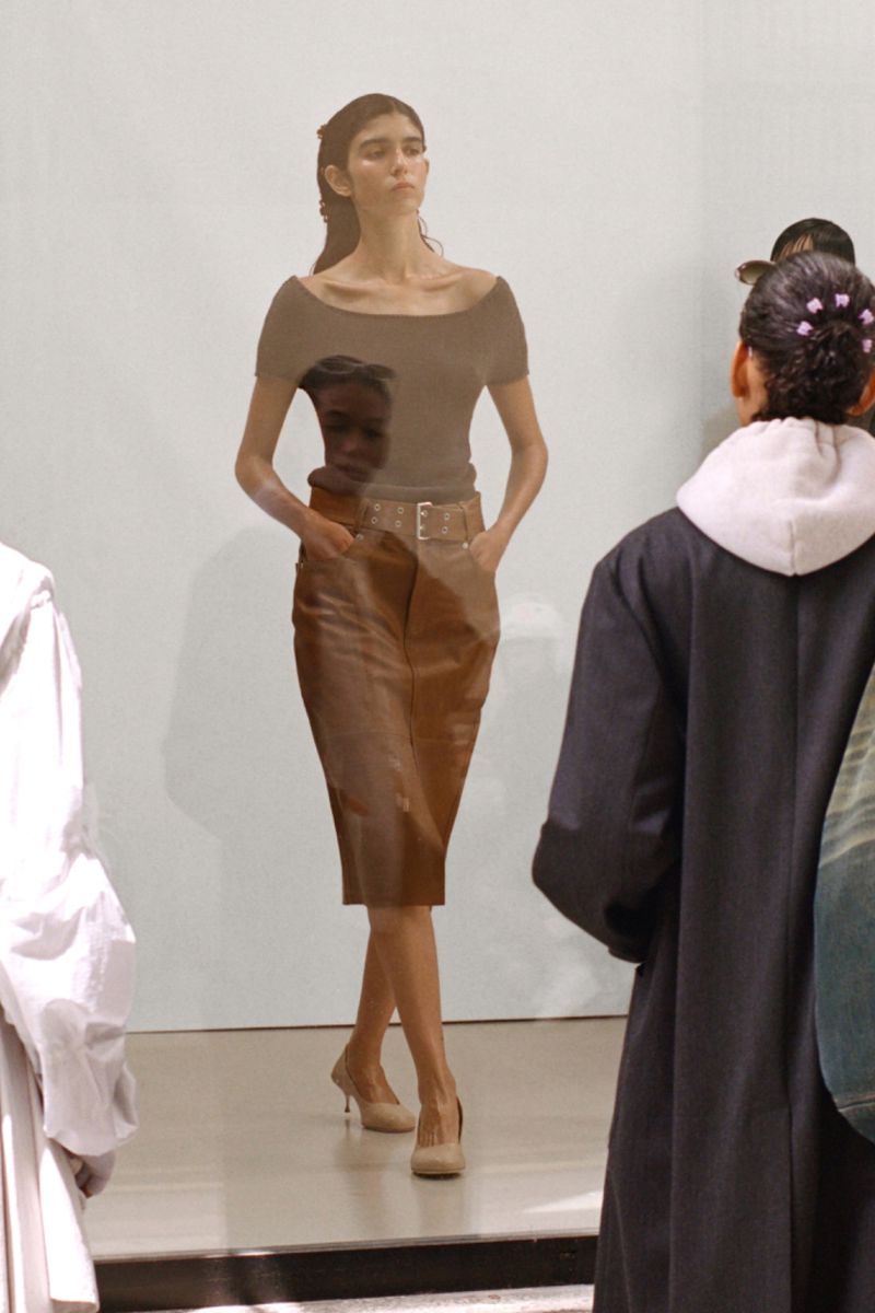 A woman walks the runway during a fashion show, wearing a brown top, tan pencil skirt with a belt, and nude heels, as an audience watches.