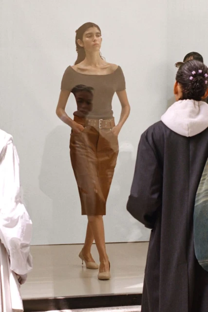 A woman walks the runway during a fashion show, wearing a brown top, tan pencil skirt with a belt, and nude heels, as an audience watches.