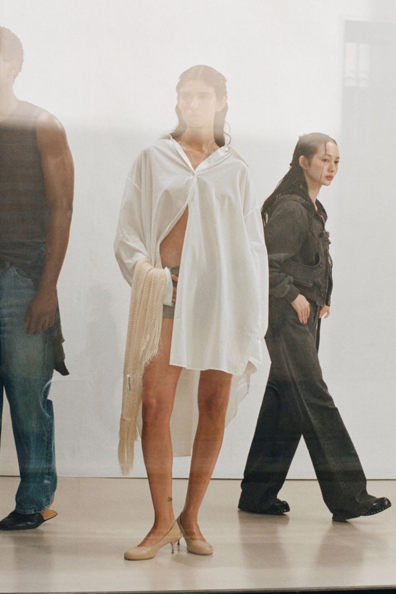Fashion editorial featuring a woman in a white shirt dress and heels flanked by a man and woman on a light studio background.