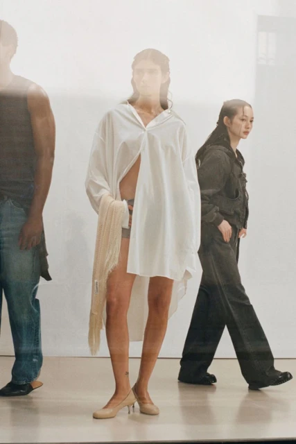 Fashion editorial featuring a woman in a white shirt dress and heels flanked by a man and woman on a light studio background.