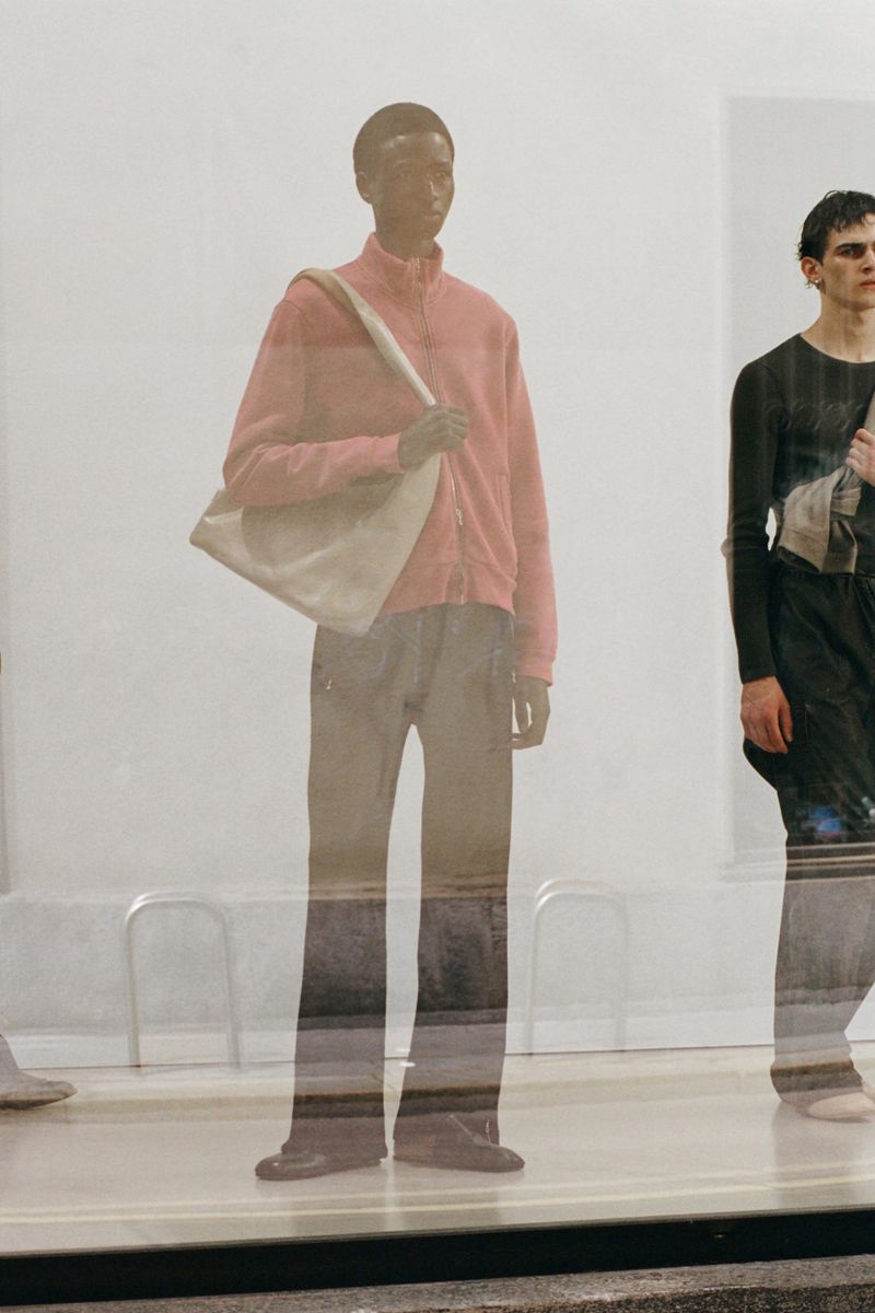 Two fashion models in studio. One wears pink jacket and shoulder bag, other wears black long-sleeve shirt. White background, artificial light.