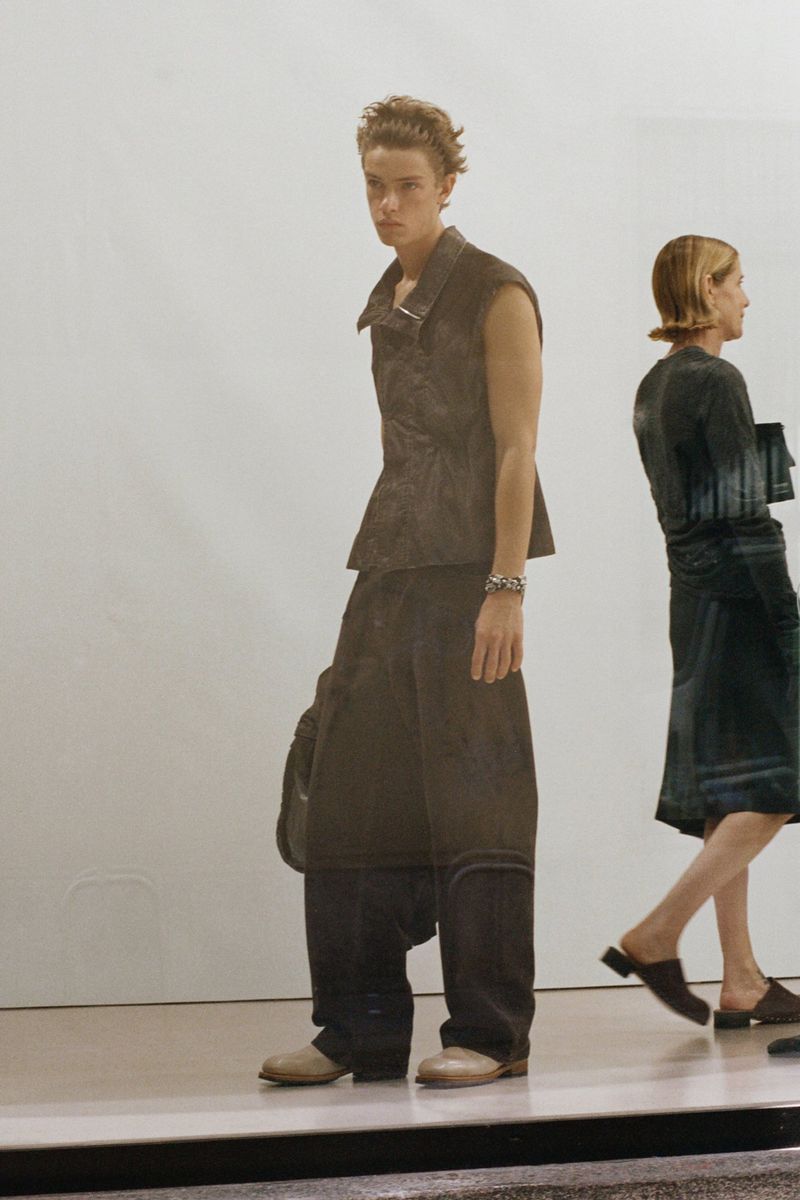 A person wearing a black vest and baggy pants stands next to another person wearing a dress, both indoors with white background.