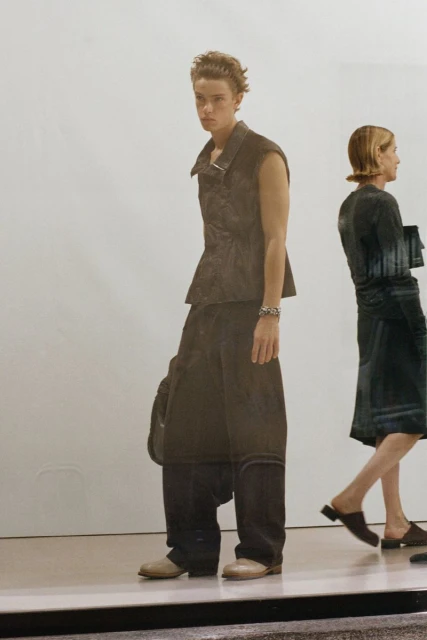 A person wearing a black vest and baggy pants stands next to another person wearing a dress, both indoors with white background.