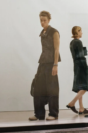 A person wearing a black vest and baggy pants stands next to another person wearing a dress, both indoors with white background.