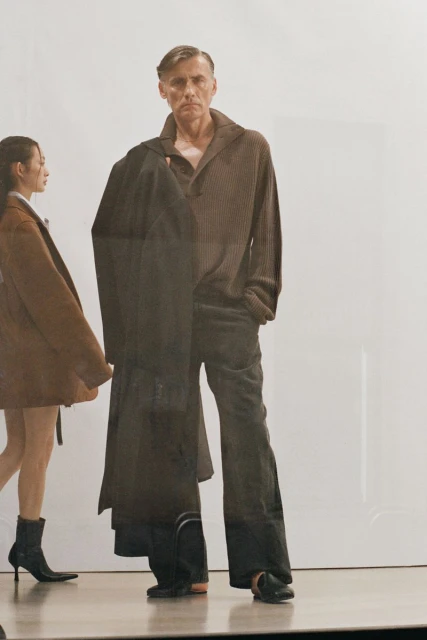 Man in brown sweater and grey pants with coat over shoulder, walking with woman in brown coat, on a reflective floor in studio.