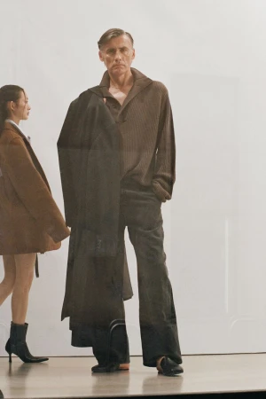 Man in brown sweater and grey pants with coat over shoulder, walking with woman in brown coat, on a reflective floor in studio.