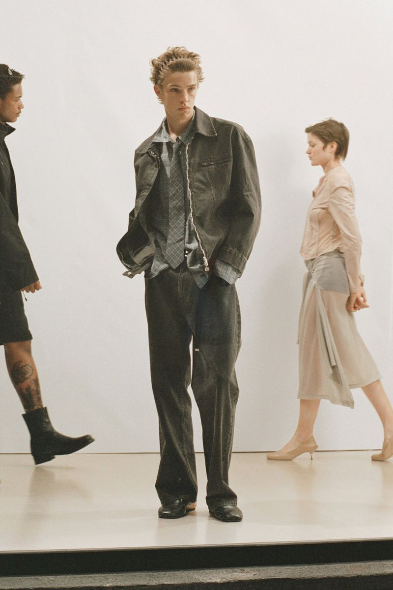 Fashion shot of a young man in a jacket and tie, with two other people in the background against a white wall in a photo studio.