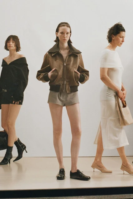 Three fashion models in a photo studio, one in a brown jacket and shorts, one in a black dress, and one in a white dress.