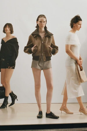 Three fashion models in a photo studio, one in a brown jacket and shorts, one in a black dress, and one in a white dress.