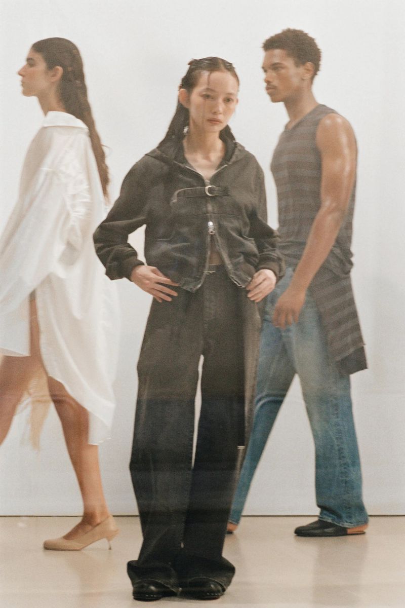 Three fashion models in an indoor setting, one wearing a black denim outfit, another a white dress, and the third a gray top and jeans.