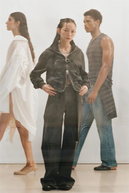 Three fashion models in an indoor setting, one wearing a black denim outfit, another a white dress, and the third a gray top and jeans.