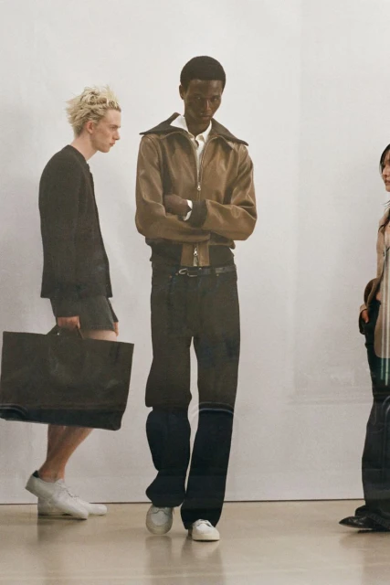Fashion shot featuring a Black male model in a brown jacket and dark pants, alongside two other models on a light background.