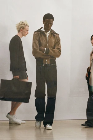 Fashion shot featuring a Black male model in a brown jacket and dark pants, alongside two other models on a light background.