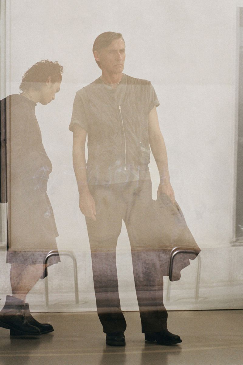 Two figures in an indoor setting, one walking with head down, the other standing upright in a casual top and trousers, looking to the right.