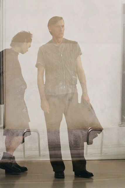 Two figures in an indoor setting, one walking with head down, the other standing upright in a casual top and trousers, looking to the right.