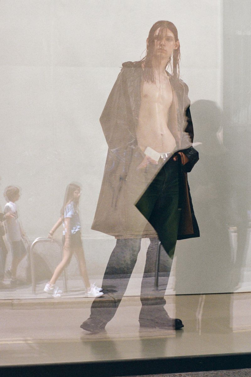 Fashion shot featuring a long-haired adult male partially nude and wearing a beige long coat, with a faint cityscape double-exposure.