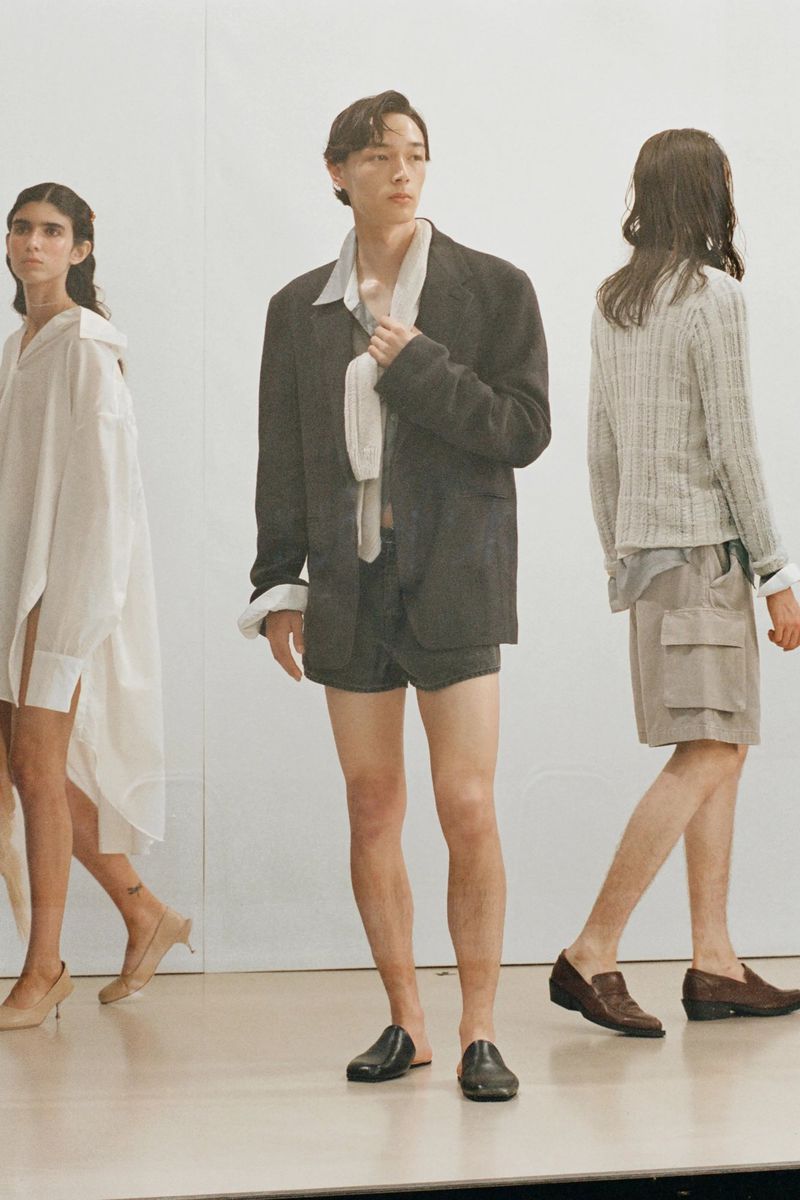 Three models pose in a minimalist fashion shoot. The central figure wears a black blazer and shorts, complemented by two others in neutral outfits.