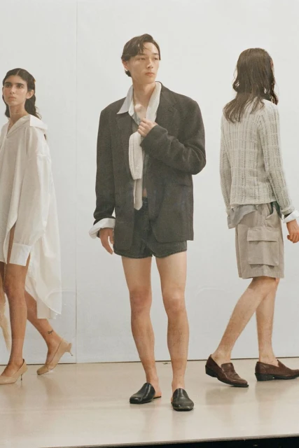 Three models pose in a minimalist fashion shoot. The central figure wears a black blazer and shorts, complemented by two others in neutral outfits.