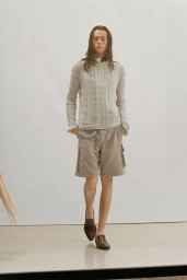 Model with long dark hair wearing a patterned knit top and cargo shorts walking forward against a white seamless background.