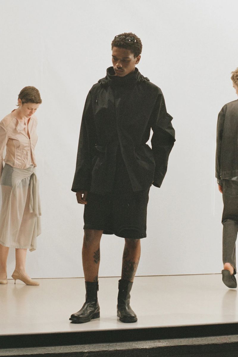 Black man wearing dark hooded jacket, shorts, and boots standing centrally with other figures against a white wall.