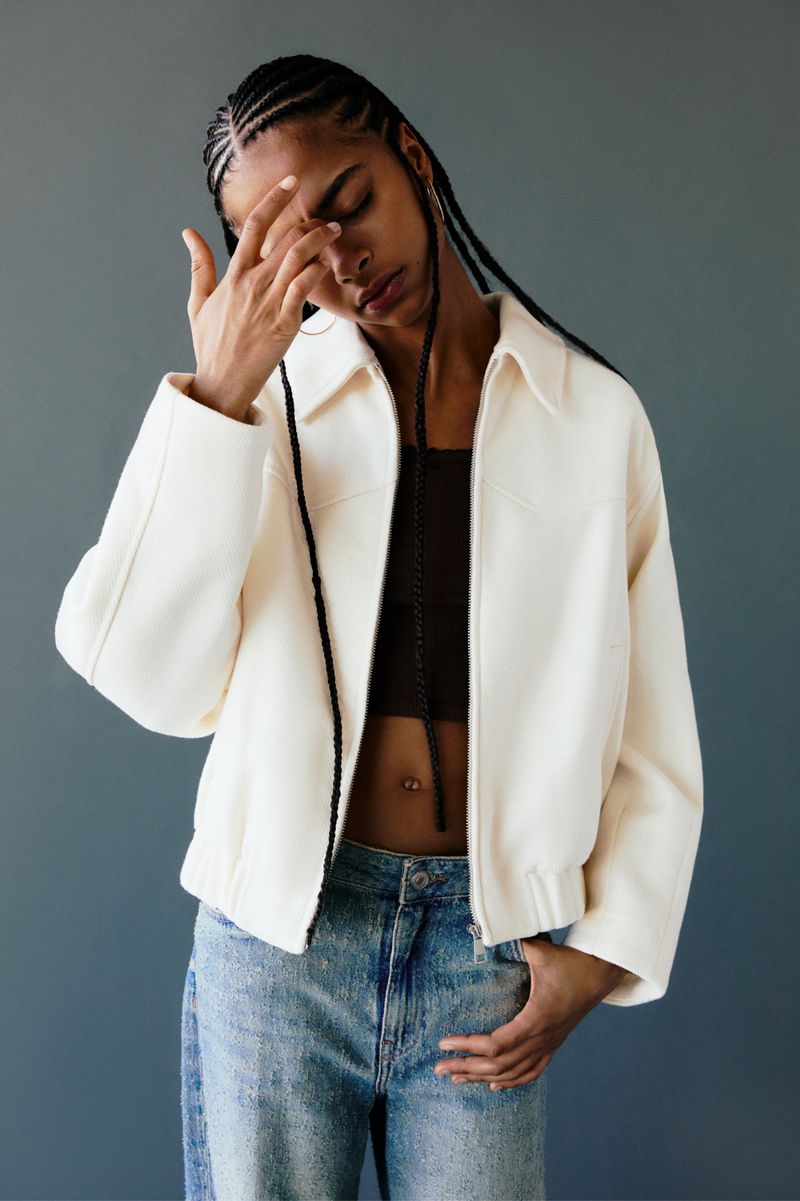 Woman in a white zip-up jacket and blue jeans with her hand to her forehead against a grey studio backdrop.