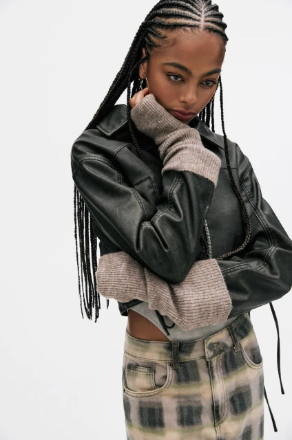 Young woman in black leather jacket and plaid denim skirt. She has black braided hair and is posing against a white background.