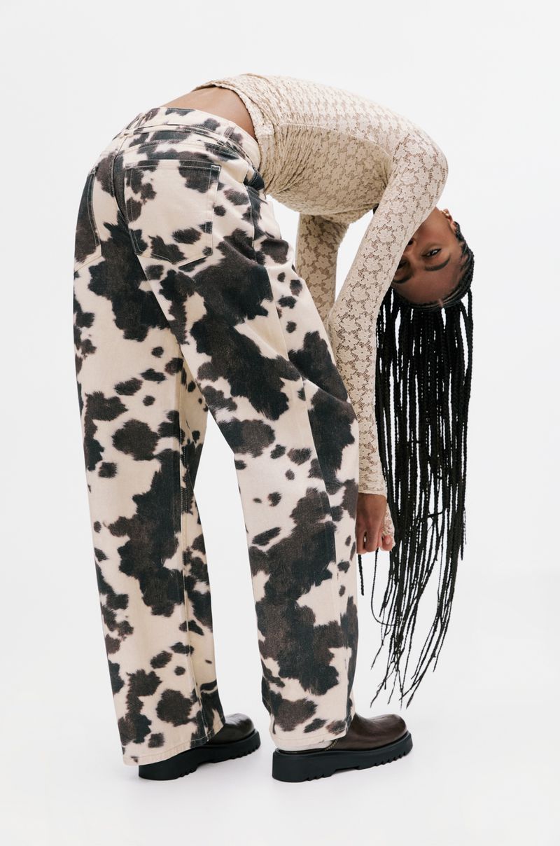 Woman in a patterned top and cow-print jeans bends over in a white photo studio, with brown shoes and long braided hair.