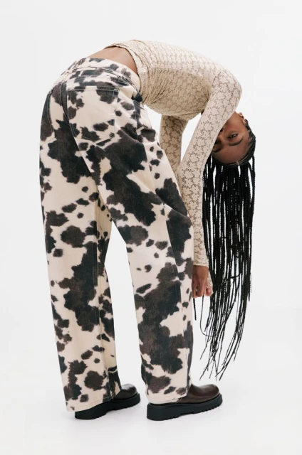 Woman in a patterned top and cow-print jeans bends over in a white photo studio, with brown shoes and long braided hair.
