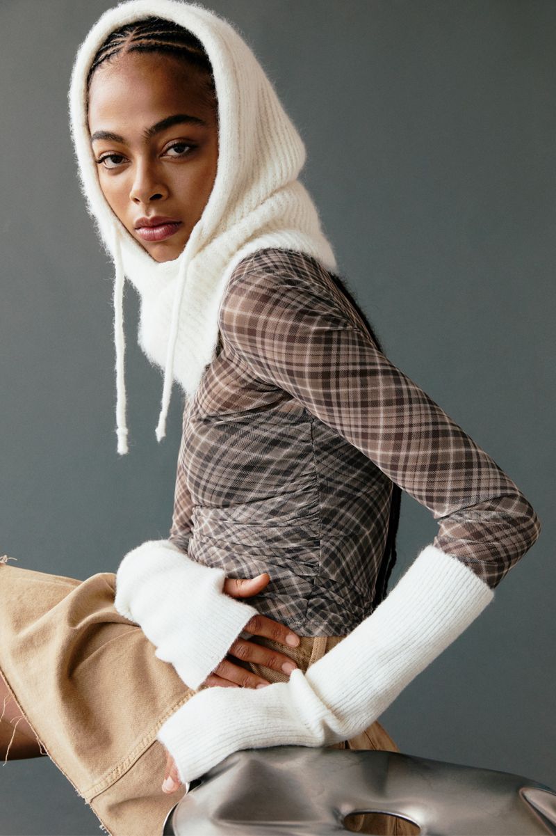 Portrait of a stylish African-American woman wearing a knit balaclava and plaid top while seated against a grey backdrop in a studio.
