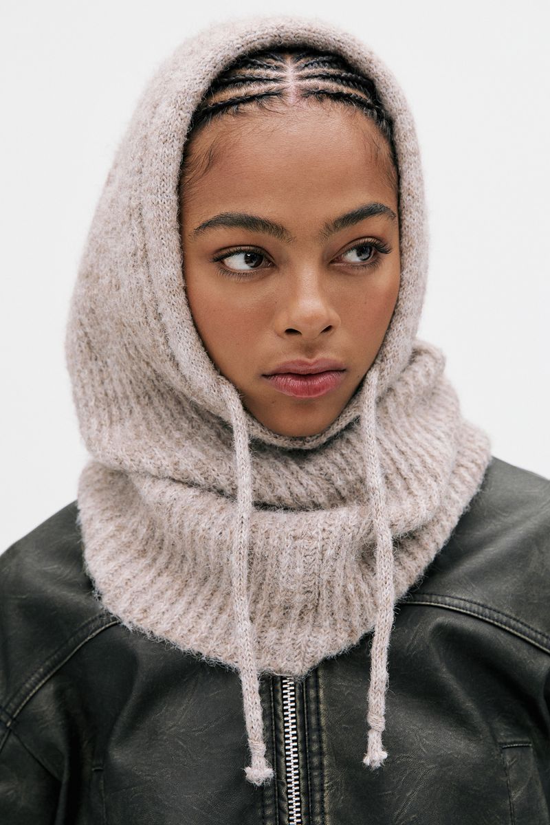 A woman poses indoors wearing a knitted balaclava with drawstrings and a black leather jacket against a white background in a studio shot.